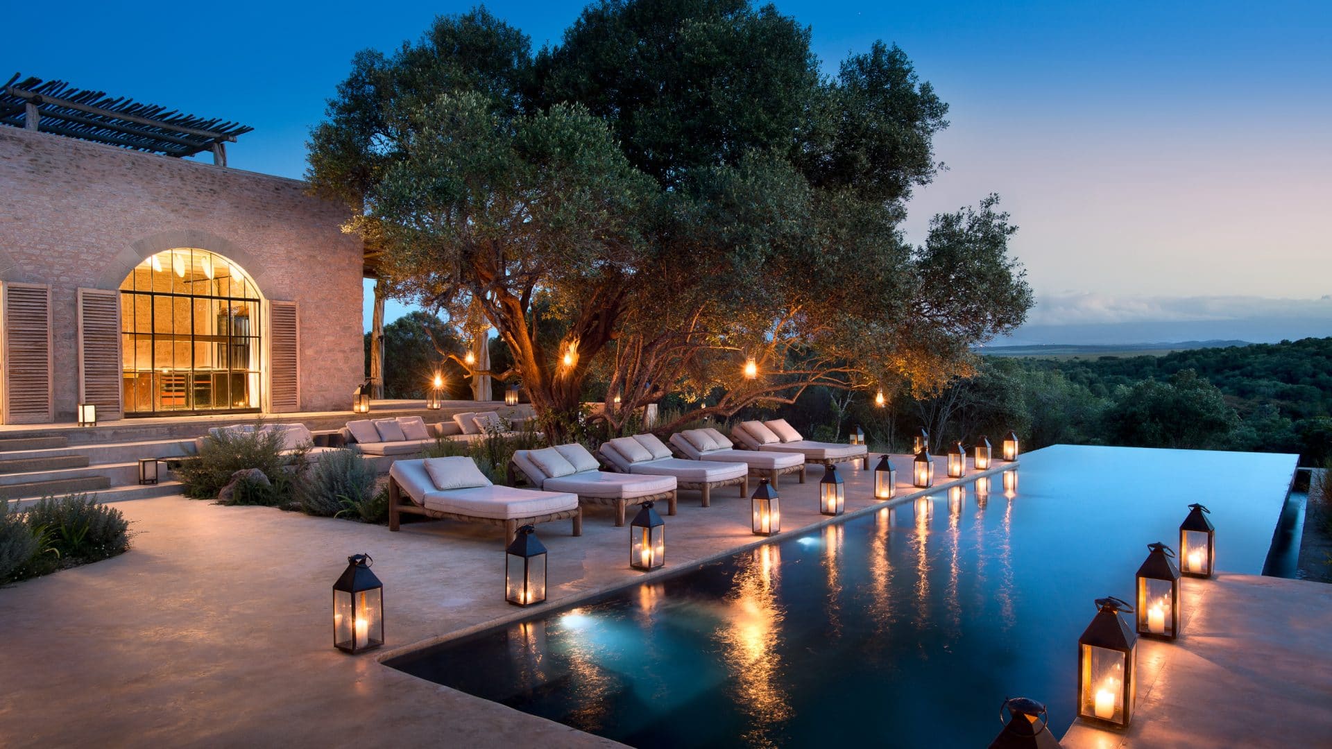A luxury lodge terrace at dusk with candle lanterns lining an infinity pool beneath a spreading tree.