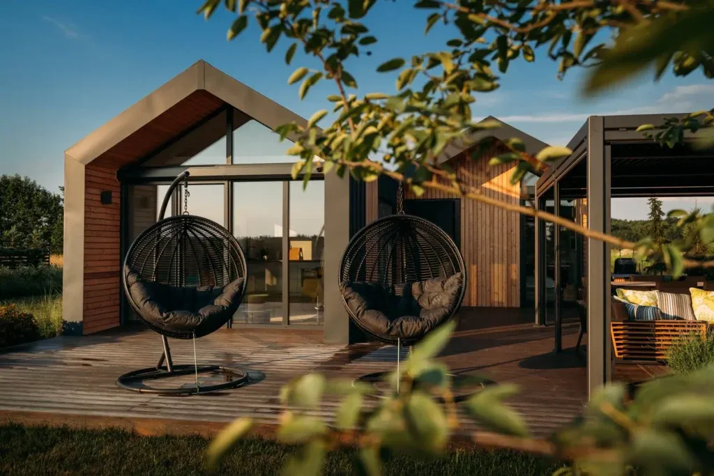 A modern timber holiday cabin with hanging egg chairs on a deck in golden evening light.